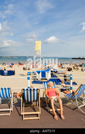 A female asleep on a deckchair on the promenade in front of the crowded sandy beach at Weymouth. Stockfoto
