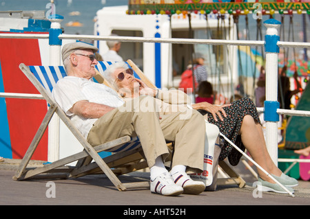 Close up of an elderly couple pensioners on deckchairs on the promenade in front of the crowded sandy beach at Weymouth. Stockfoto