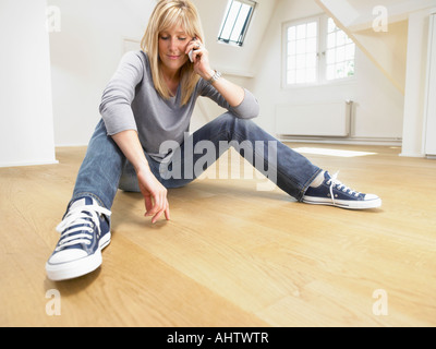 Entspannte Frau sitzend auf Holzboden geben einen Anruf mit ihrem Handy. Weißen leeren Loft. Stockfoto