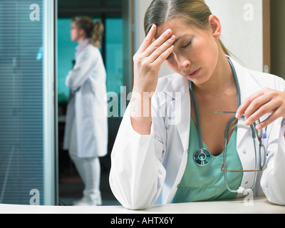 Müde Arzt sitzt an ihrem Schreibtisch mit Ärztin im Hintergrund stehen. Stockfoto