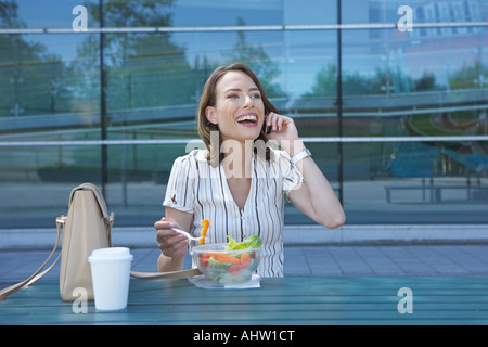 Geschäftsfrau auf Tisch Büro außerhalb auf dem Handy beim Salat essen um sich lächelnd zu suchen. Stockfoto