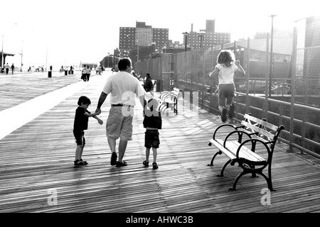 Vater und Kinder der Coney Island-Promenade schlendern, mit der jungen Mädchen-Tochter von den Bänken zu springen, wie sie Fuß Stockfoto