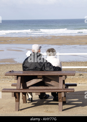 Altes Ehepaar sitzt auf einem hölzernen Picknickbank am Crooklets Strand, mit Blick auf das Meer. Stockfoto