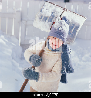 Lächelndes Kind greift ein Snowshovel in seinem blauen stricken Handschuhe, steht er außerhalb an einem verschneiten Sonnentag Stockfoto