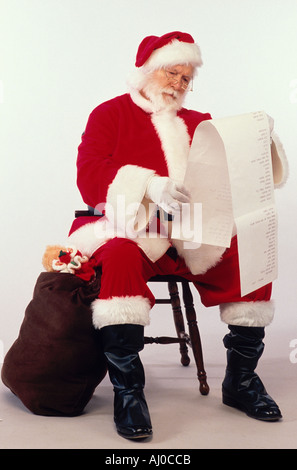 Santa Claus berät eine lange Liste von Anforderungen, wie er auf einem Hocker neben seiner Tasche von Spielzeug sitzt Stockfoto