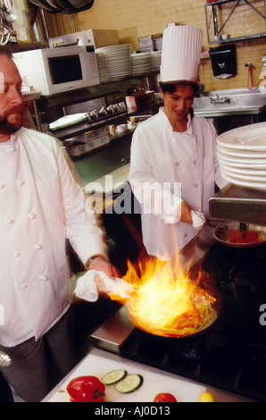 Zwei Köche kochen auf dem Herd in einer Restaurantküche eines ihre Pfannen ist absichtlich in Flammen Stockfoto