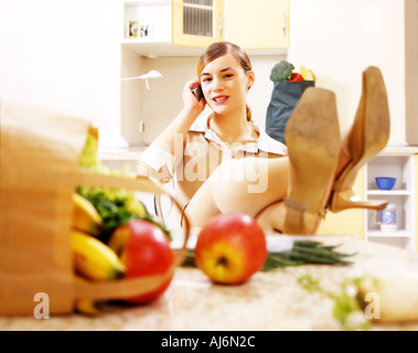 Frau in der Küche mit ihren Beinen auf dem Tisch sitzen und telefonieren. Stockfoto