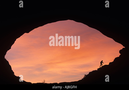 USA Utah Arches National Park Wanderer steigt am Rand des natürlichen Bogen als Sonnenaufgang Lichter hohe Wolken im Morgengrauen Stockfoto