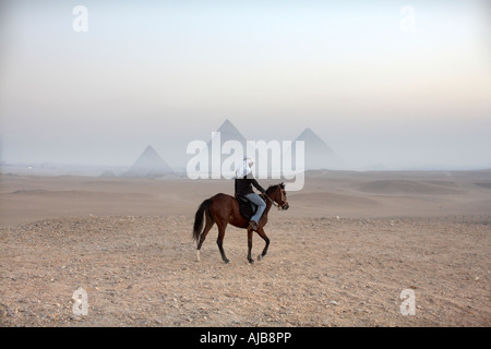 Pferd Reiter Guide Reiten in der Steinwüste in am frühen Morgendämmerung dunstigen Nebel mit Pyramiden darüber hinaus in Ferne Gizeh Kairo Ägypten Stockfoto