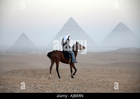 Pferd Reiter Guide Reiten in der Steinwüste mit entfernten Pyramiden jenseits im frühen Morgengrauen dunstigen Morgennebel in Gizeh Kairo Ägypten A Stockfoto