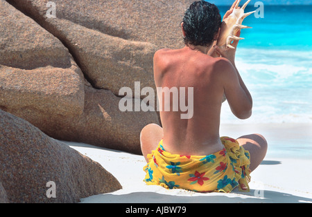 Frau sitzt zwischen Felsen am Strand, gelbes Tuch tragen, halten Schale an sein Ohr Stockfoto