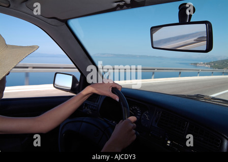 Junge Autofahrerin auf eine Küstenstraße mit dem Meer und blauer Himmel sichtbar durch die Auto-Windschutzscheibe Stockfoto