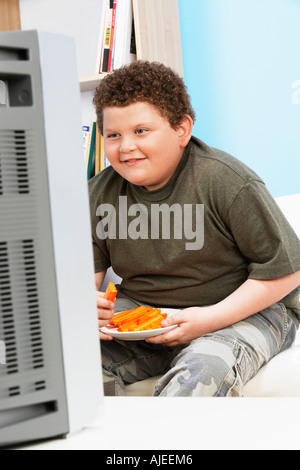 Übergewichtige junge klebt (13-15) Essen Karotte vor Fernseher Stockfoto