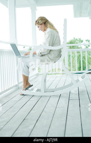 Frau sitzt im Schaukelstuhl auf der Veranda, mit Laptop-Computer, in voller Länge Stockfoto