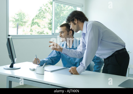 Geschäftsmann, sitzen am Schreibtisch vor Monitor, während Kollege neben ihm, zu sprechen und zeigt auf dem Bildschirm steht Stockfoto