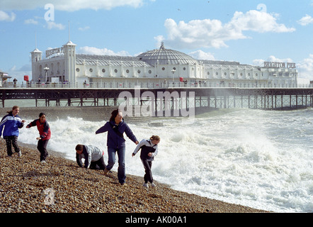 Menschen, die von den Wellen am Strand mit dem Brighton Pier im Hintergrund laufen Stockfoto