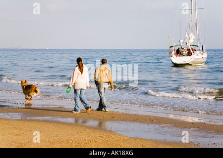 Ein paar Spaziergang mit ihrem Hund am Strand von Bournemouth, Dorset. VEREINIGTES KÖNIGREICH.  Und eine Yacht vor Anker in der Nähe der Küste. Stockfoto