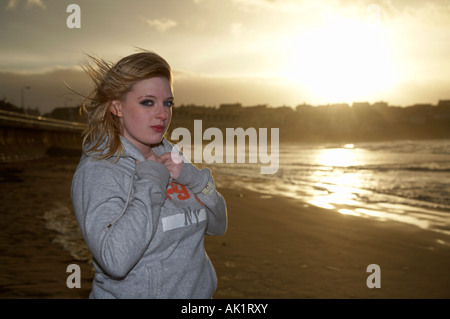 blonde kurzhaarige Studentin mit Tränen in den Augen stehen am Strand bei Sonnenuntergang eine Sweatshirt hielt ihn am Hals zu tragen Stockfoto