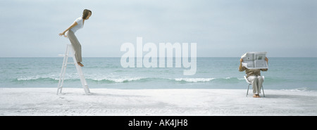 Frau auf Leiter am Strand, Mann liest Zeitung Stockfoto