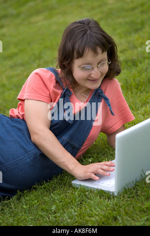 FRAU, LÄCHELN, ARBEITEN UND LERNEN VOR EINEM LAPTOPCOMPUTER, AUF DEM RASEN DRAUßEN IM PARK Stockfoto