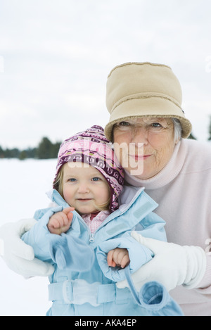 Senior Woman umarmen Kleinkind Mädchen, beide Lächeln, gekleidet in Winterkleidung, Porträt Stockfoto