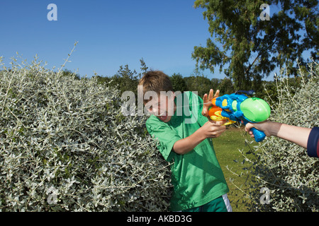 Zwei Jungs (6-11) spielen mit Wasserpistolen unter Büschen, lachen, close-up von Hand mit Pistole Stockfoto