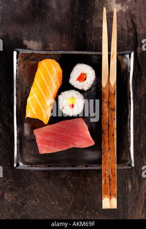 SCHWARZES QUADRAT-PLATTE MIT ZWEI STÜCKE VON ROHEM FISCH UND ZWEI KREISE VON VERPACKTEN REIS HÖLZERNE STÄBCHEN SCHWARZ TISCHPLATTE Stockfoto