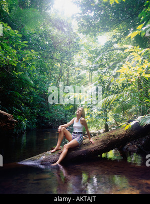 Frau, die auf einem umgestürzten Baumstamm über einem Bach in einem üppig grünen Wald sitzt, Sonnenlicht durchs Laub filtert Stockfoto