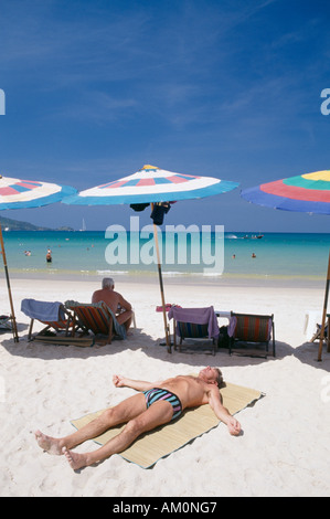 THAILAND Phuket Patong Beach Sonnenanbeter liegen auf dem Rücken im Vordergrund mit Reihe von Sonnenschirmen und Liegestühlen. Mann zum Sonnenbaden. Stockfoto