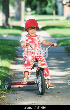 Junges Mädchen mit rotem Hut und gestreifte Overalls Dreirad fahren Stockfoto