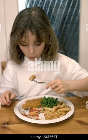 sieben Jahre altes Mädchen essen Fischstäbchen Pommes Frites und Erbsen am Tisch Stockfoto