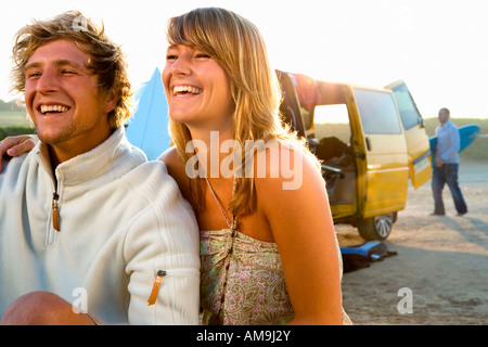 Paar am Strand lächelnd mit Mann mit Surfbrett im Hintergrund sitzt. Stockfoto