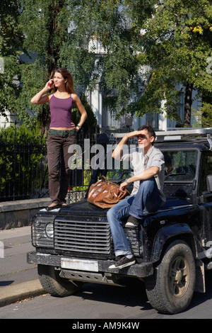 Frau stehend auf Motorhaube des SUV auf Handy mit Mann sitzt auf der Haube. Stockfoto