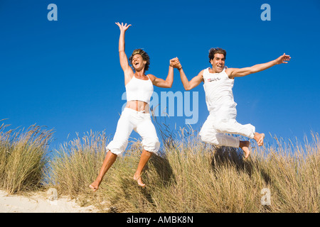 junges Paar Hand in Hand am Strand mit erhobenen Armen springen Stockfoto
