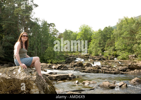 Junge Frau sitzt auf einem Felsen durch die Falls of Dochart in Killin Perthshire Schottland Stockfoto