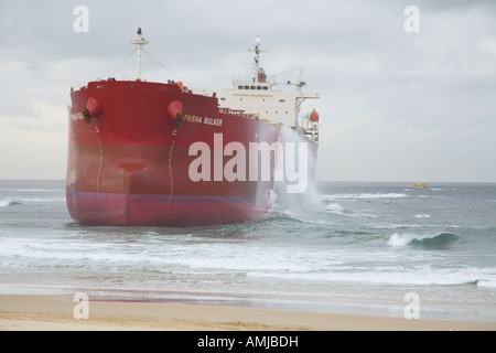 Großes rotes Schiff, das am Sandstrand gestrandet ist, mit Wellen, die unter dem bewölkten Himmel gegen den Rumpf stürzten Stockfoto