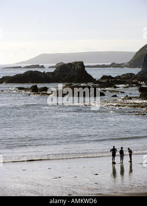 Drei silhouetted Zahlen über Ayrmer Cove Beach mit felsigen Klippen und einen glitzernden Meer in silbrig, am späten Nachmittag Sonne. South Devon, Großbritannien Stockfoto