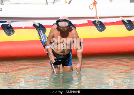 Alter Mann mit Badeshorts im seichten Meer mit einem Metalldetektor wasserdicht für Elemente verloren durch Urlauber suchen. Stockfoto