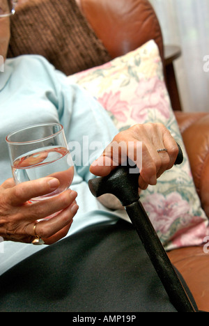 Eine ältere Frau sitzt bequem auf einer Couch, stützt sich mit einem Stock in der einen Hand und hält ein Glas Wasser in der anderen Stockfoto
