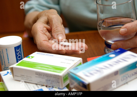 Hände einer älteren Frau hält eine Pille und ein Glas Wasser umgeben von Kisten mit ihrem verschreibungspflichtige Medikamente Stockfoto