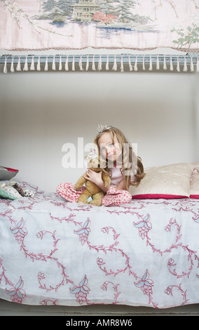 Mädchen auf dem Bett sitzend, hält Teddybär und trägt Tiara Stockfoto