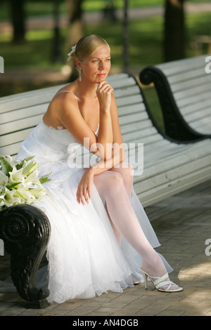 Eine nachdenkliche Braut in einem weißen Kleid sitzt auf einer Parkbank Stockfoto