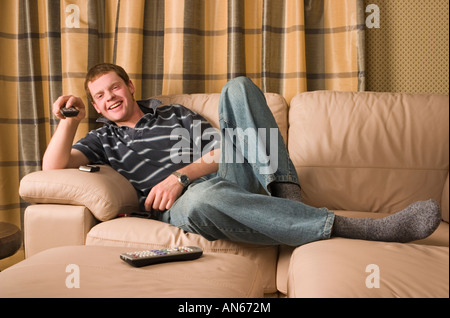 Mann in seinen Zwanzigern, Lächeln, eine TV-Fernbedienung zu halten, während Sie auf dem Sofa entspannen Stockfoto