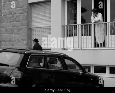 Frau in weißen Handtuch Bademantel stützte sich auf ihre Balkongeländer gerade einen Mann trägt einen schwarzen Hut zu Fuß durch in der französischen Provinz Stockfoto