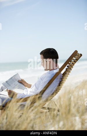 Mann sitzt im Liegestuhl am Strand, lesen Zeitung Stockfoto