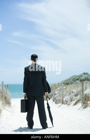 Geschäftsmann am sandigen Weg zum Ozean, holding, Aktenkoffer und Sonnenschirm, Rückansicht Stockfoto