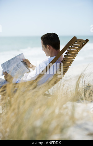 Junger Mann sitzt im Liegestuhl am Strand, lesen Zeitung Stockfoto