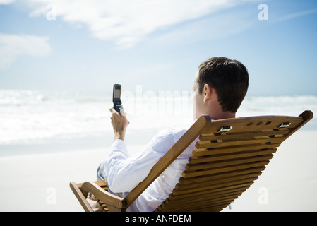 Mann sitzt im Stuhl am Strand, mit Handy, Rückansicht Stockfoto