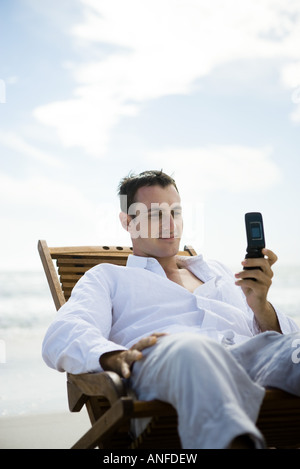 Junger Mann sitzt im Stuhl am Strand, mit Handy Stockfoto