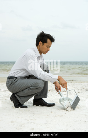 Mann kauert auf Strand mit Besen und Kehrschaufel Stockfoto